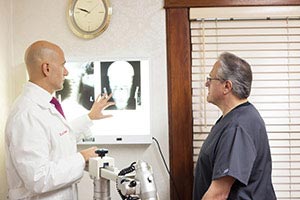 The image shows a medical professional presenting a patient s X-ray to another person in a room with a window and blinds, while two other individuals observe.
