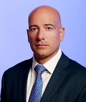 The image features a man wearing a suit and tie, posing with a professional expression against a blue background.