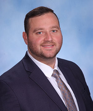 The image shows a man standing against a blue background with a school logo on the wall behind him. He appears to be wearing a suit and tie, smiling slightly towards the camera.