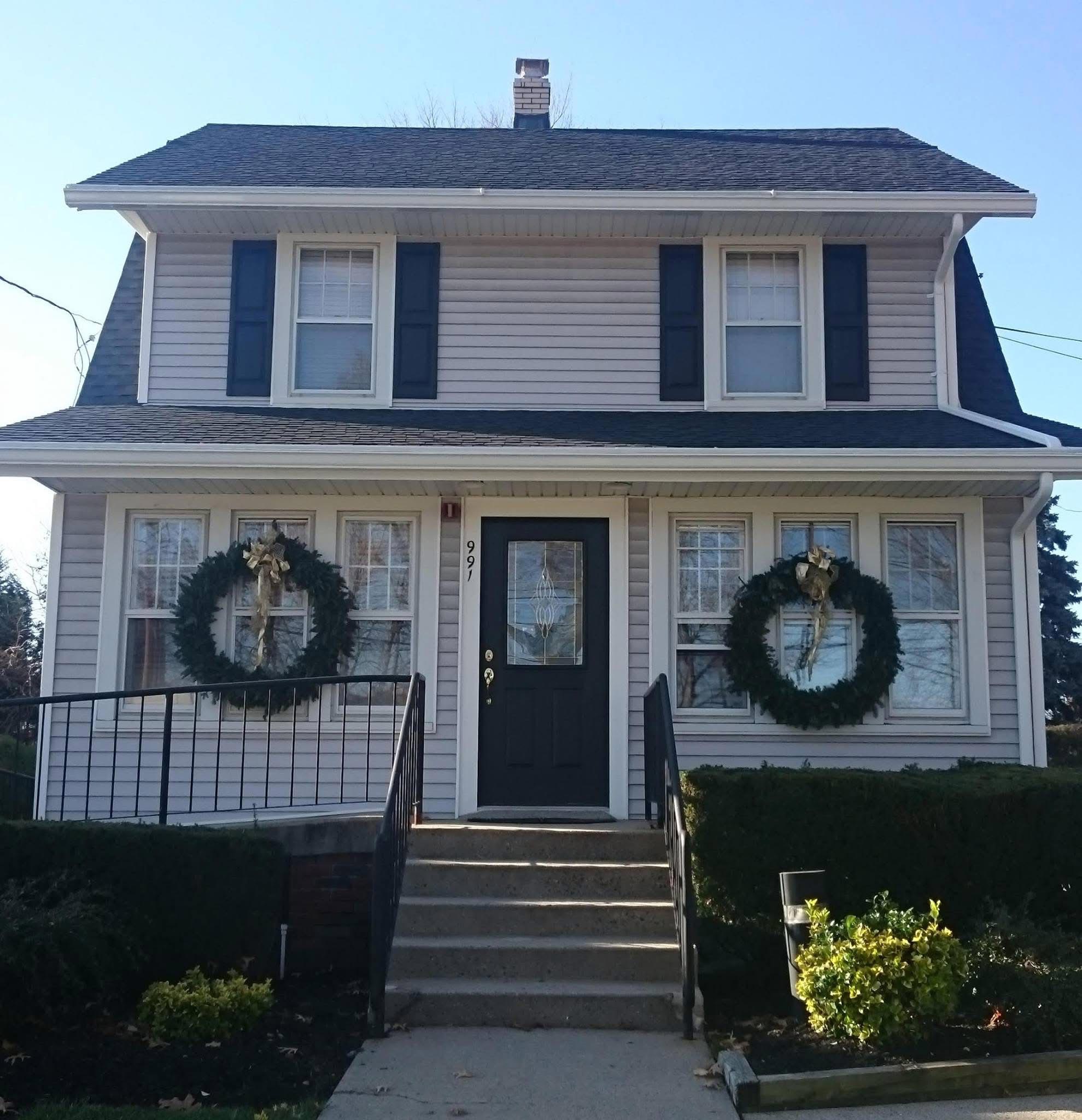 The image shows a two-story house decorated with Christmas wreaths on the front door and windows, adorned with seasonal lights, and featuring a black roof and white trim.