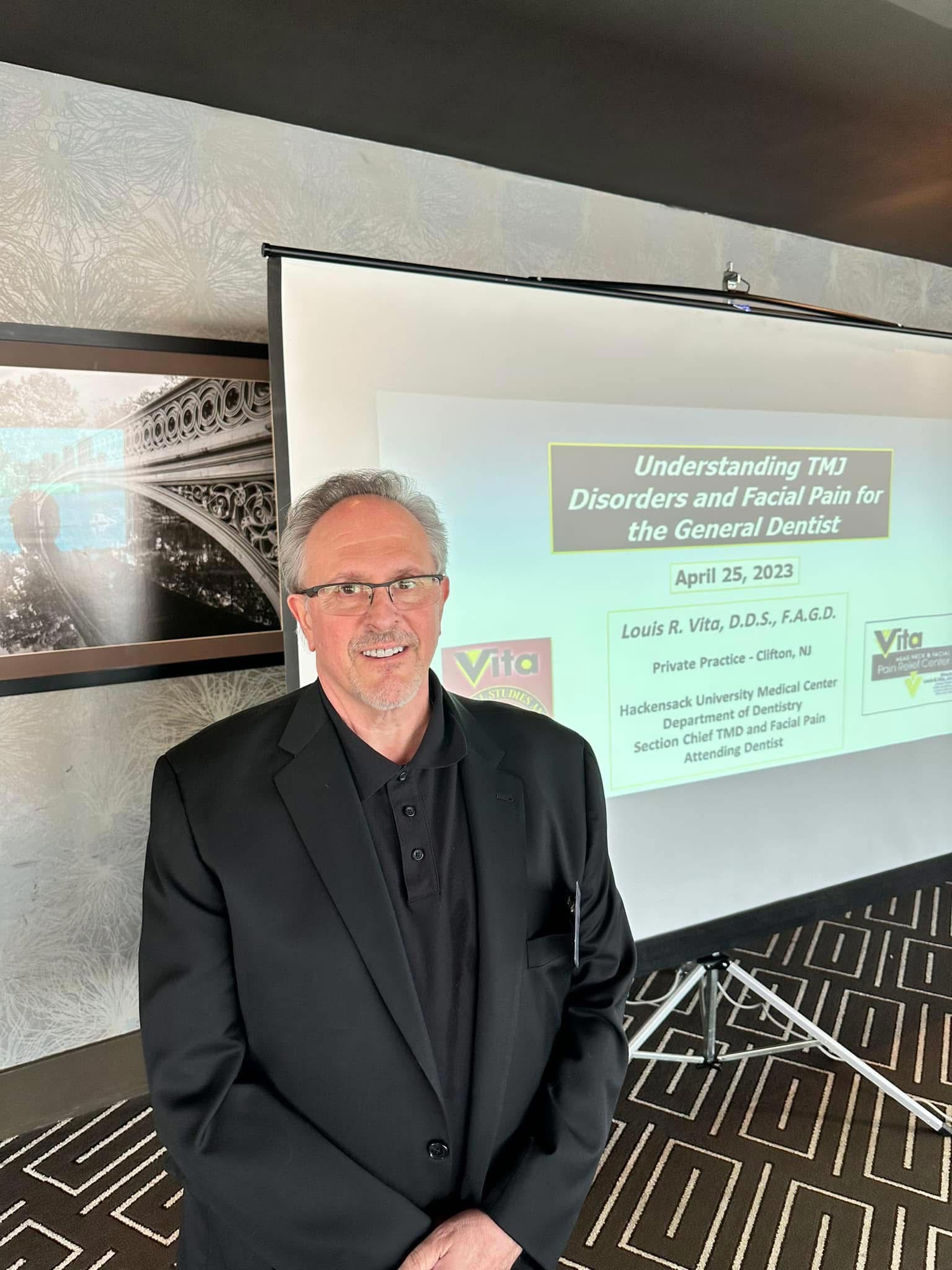 The image shows a man standing at the front of a presentation, with a slide displaying information about understanding TMJ disorders and facial pain. He appears to be a presenter or speaker, addressing an audience.