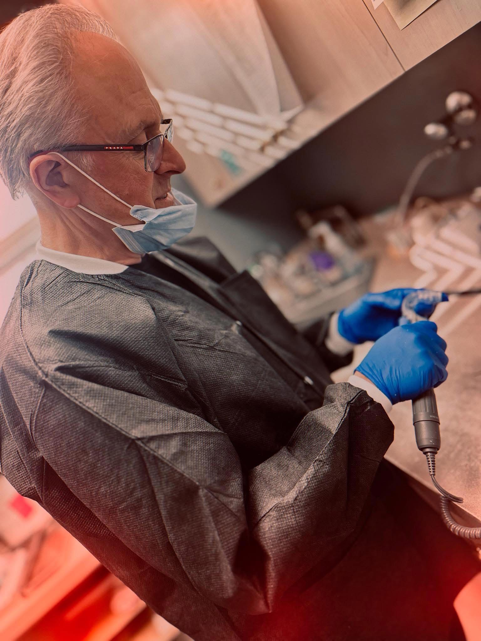 The image features a man wearing a face mask and gloves, standing in a laboratory setting with various scientific equipment around him, focusing on a microscope.