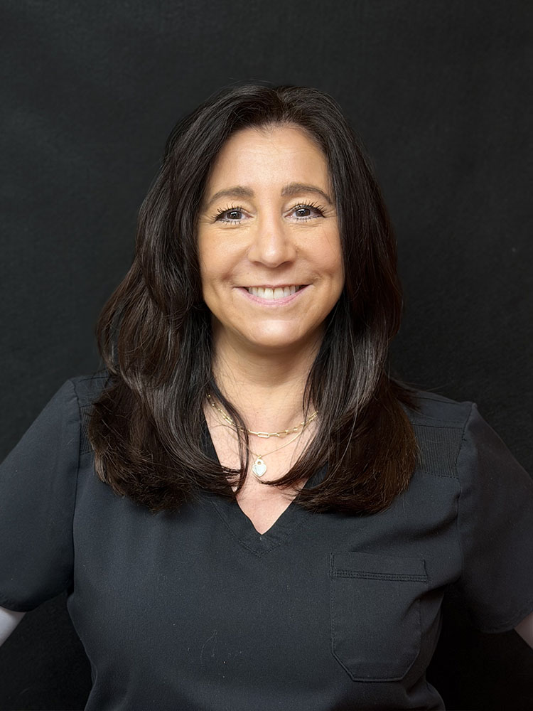 This is a color photograph of a woman with short brown hair, wearing a dark top, standing against a black background.