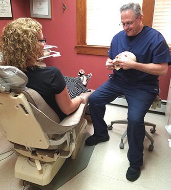 The image shows a man sitting in a dental chair with his mouth open, while a woman stands beside him holding a small animal. They are in an office setting with dental equipment visible.