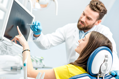 A man in a white coat stands next to a woman in a dental chair, both in a dental office setting, with a large screen displaying medical images behind them.